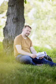 Janne Ripatti istuu puun alla ruohikolla kirja kädessään.