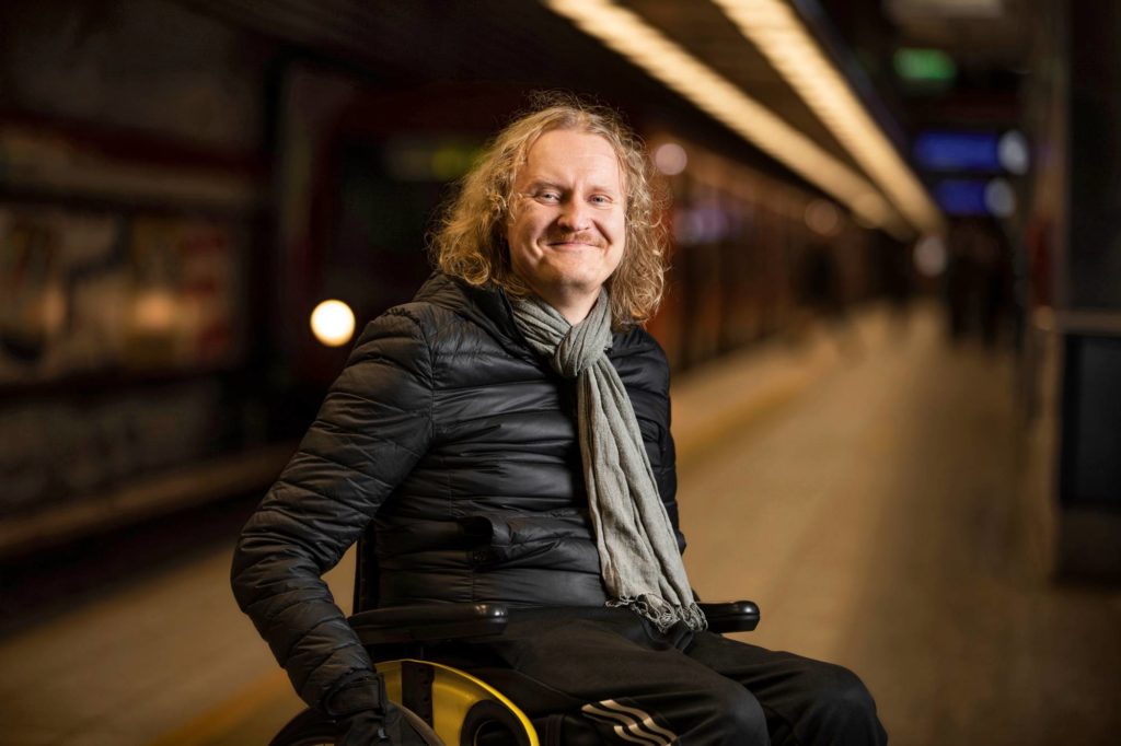 Tuomas Hakamäki on kuvattu metroasemalla. Kuvassa hän istuu hymyillen pyörätuolissa metrojunan edessä.