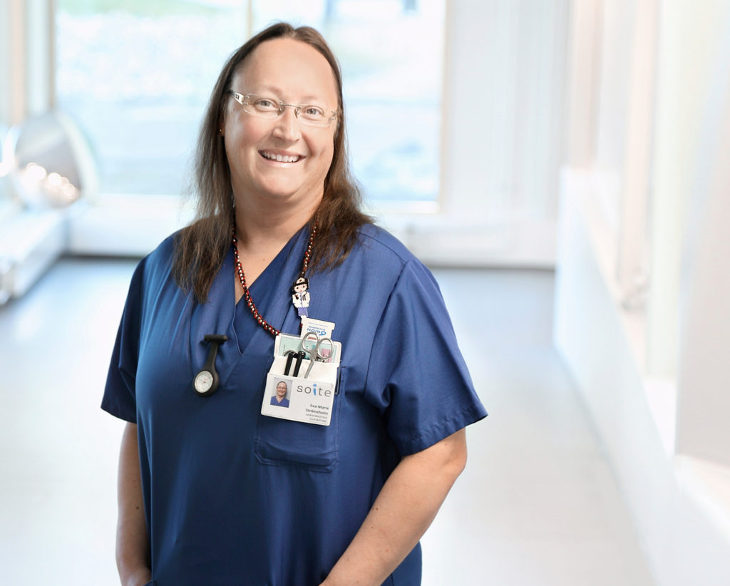 Eva-Maria Strömsholm seisoo hymyillen sairaanhoitajan työvaatteissa osaston käytävällä.